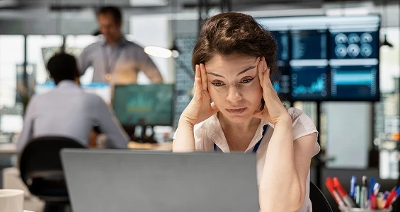 Zmartwiona i sfrustrowana kobieta w biurze trzymająca się za głowę przed laptopem, sugerująca chaos informacyjny i potrzebę wdrożenia systemu BPMS.
