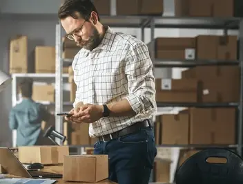 Pracownik magazynu z telefonem przy biurku z paczką i laptopem, w tle regały z przesyłkami - automatyzacja procesów e-commerce i zarządzanie zamówieniami.