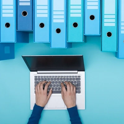 Widziany od góry dłonie pracujące na laptopie i segregatory jako symbol elektronicznych teczek pracowników
