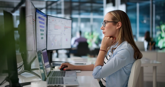 Kobieta w okularach pracuje przy biurku w biurze, analizując dane na kilku monitorach i laptopie