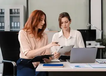 Dwie kobiety w strojach biznesowych siedzą przy biurku w nowoczesnym biurze i omawiają dokumenty oraz informacje na tablecie.