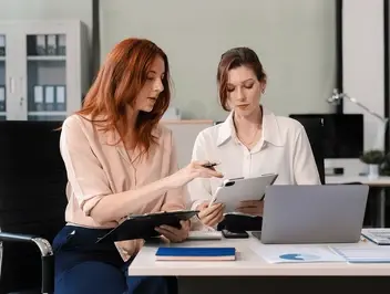 Dwie kobiety w strojach biznesowych siedzą przy biurku w nowoczesnym biurze i omawiają dokumenty oraz informacje na tablecie.