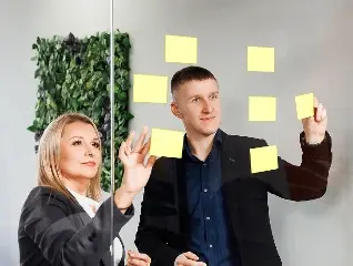 Dwoje ludzi w biznesowych strojach przy szklanej ścianie, na której przyklejone są żółte karteczki samoprzylepne. Kobieta z lewej strony patrzy na karteczki, a mężczyzna po prawej sięga po jedną z nich ręką.