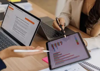 Dwie osoby przy biurku analizujące wykresy finansowe na tablecie oraz dane na ekranie laptopa.