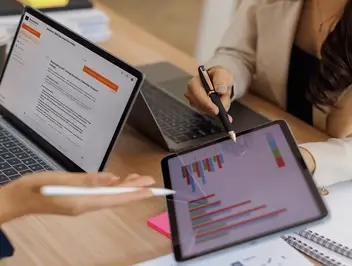 Dwie osoby przy biurku analizujące wykresy finansowe na tablecie oraz dane na ekranie laptopa.
